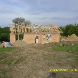 Budowa domu w stanie surowym otwartym: ceglana konstrukcja z drewnianym szkieletem dachu, worki cementu i hałda piasku na pierwszym planie, zieleń w tle.
