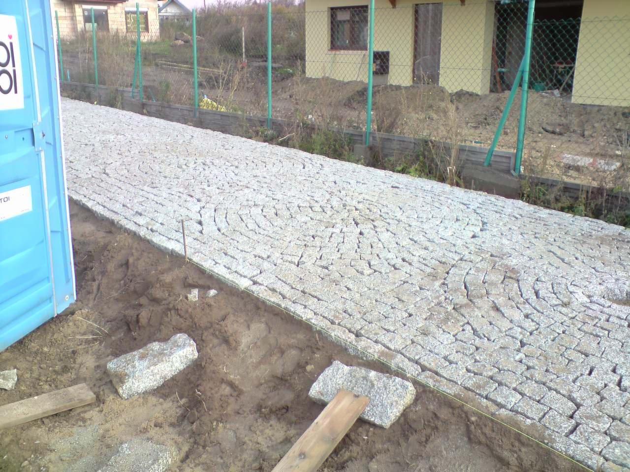 Ułożona kostka granitowa w kształcie wachlarza tworzy ścieżkę. Widoczny fragment toalety TOI-TOI i ogrodzenie z siatki.