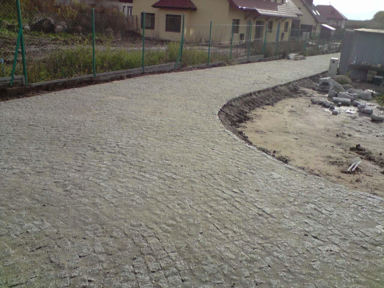 Ułożona, zakrzywiona ścieżka z kostki brukowej na podwórku, z widocznymi niedokończonymi obszarami, materiałami budowlanymi i zielonym ogrodzeniem w tle.