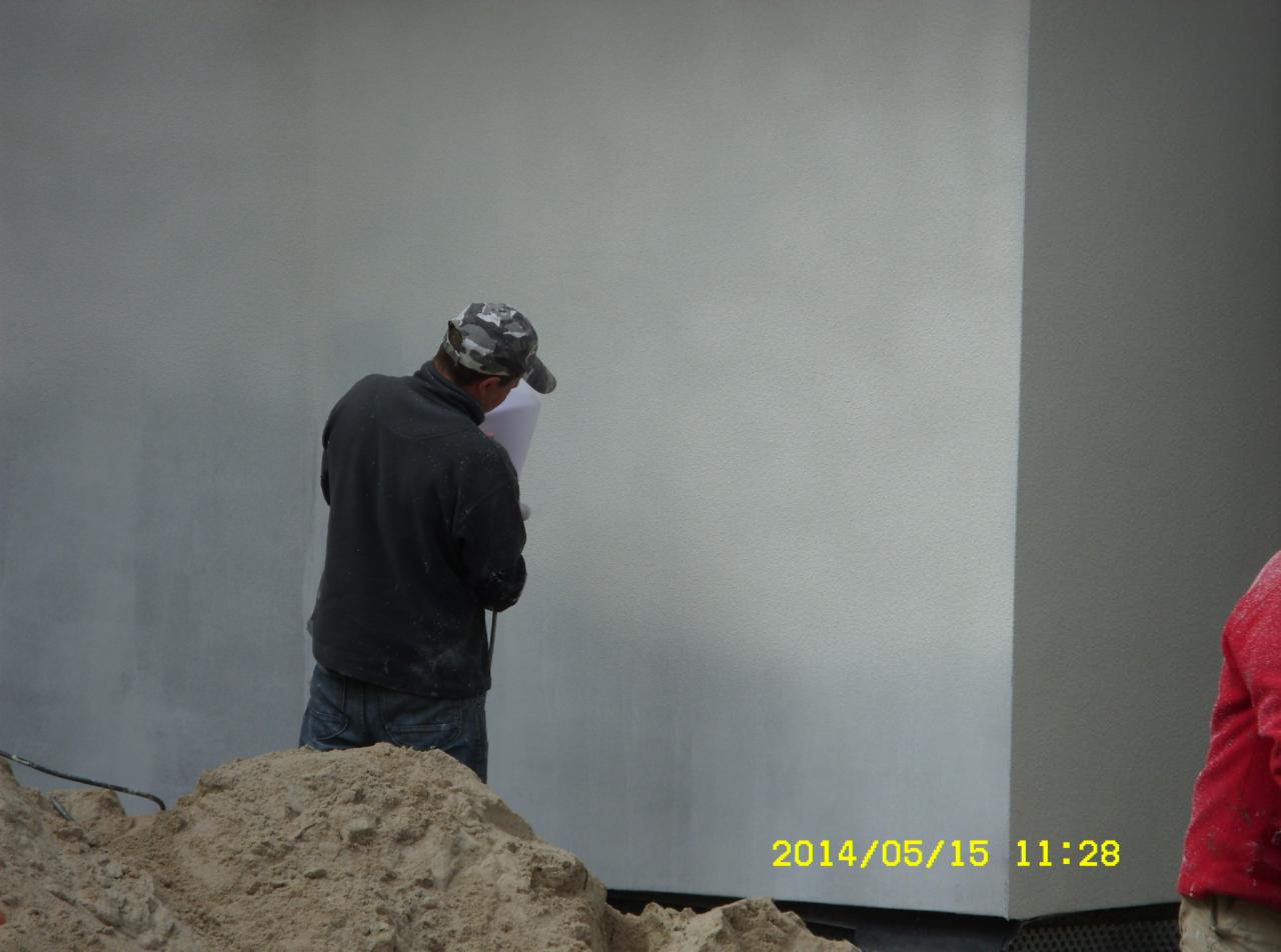 Pracownik w trakcie natrysku tynku na elewację budynku. Widoczny fragment ściany z nałożonym tynkiem, piasek i osoba w stroju roboczym. Data na zdjęciu: 2014/05/15.