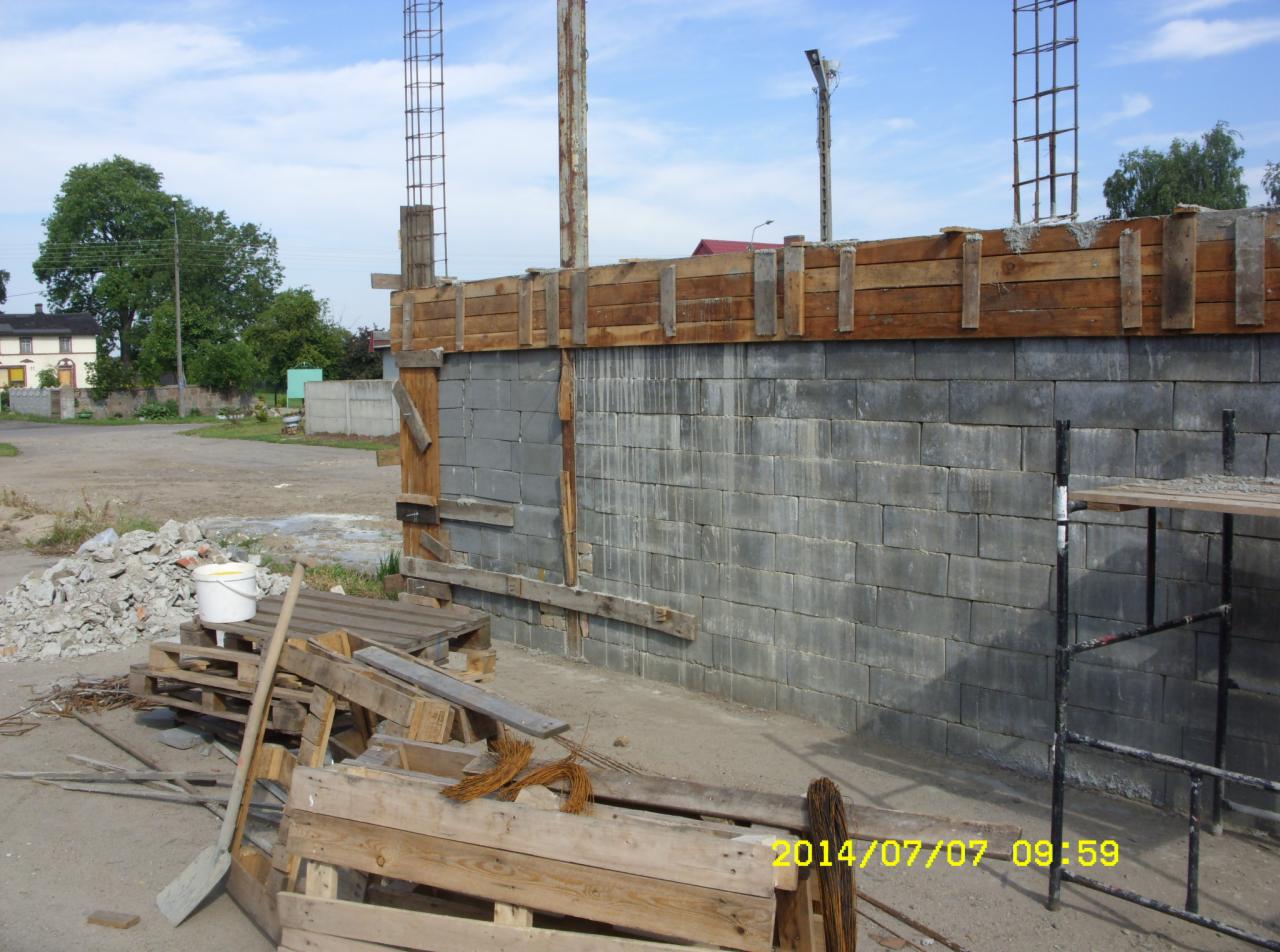 Wznoszenie fundamentów z bloczków betonowych, z widocznym szalunkiem drewnianym i zbrojeniami. Na pierwszym planie narzędzia budowlane i gruz.