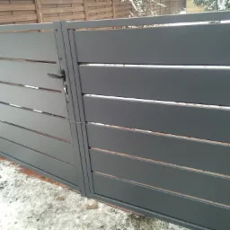 Nowoczesna, szara brama z poziomych paneli, widoczna klamka i zamek, fragment ceglanej podmurówki i śnieg.