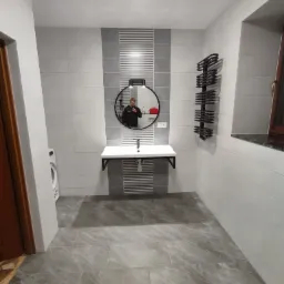 Wykończona łazienka z szarymi płytkami podłogowymi imitującymi marmur, biało-szare płytki ścienne, okrągłe lustro z podświetleniem i czarną ramą, biała umywalka na czarnych wspornikach oraz czarny...