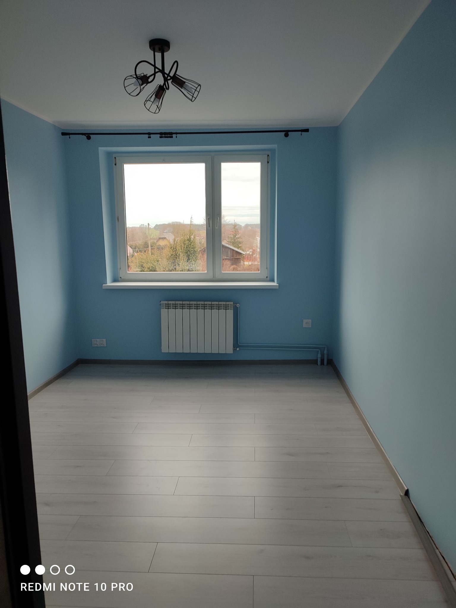 Pomalowane na błękitno puste pomieszczenie z jasną podłogą, grzejnikiem pod oknem i nowoczesną lampą sufitową.
