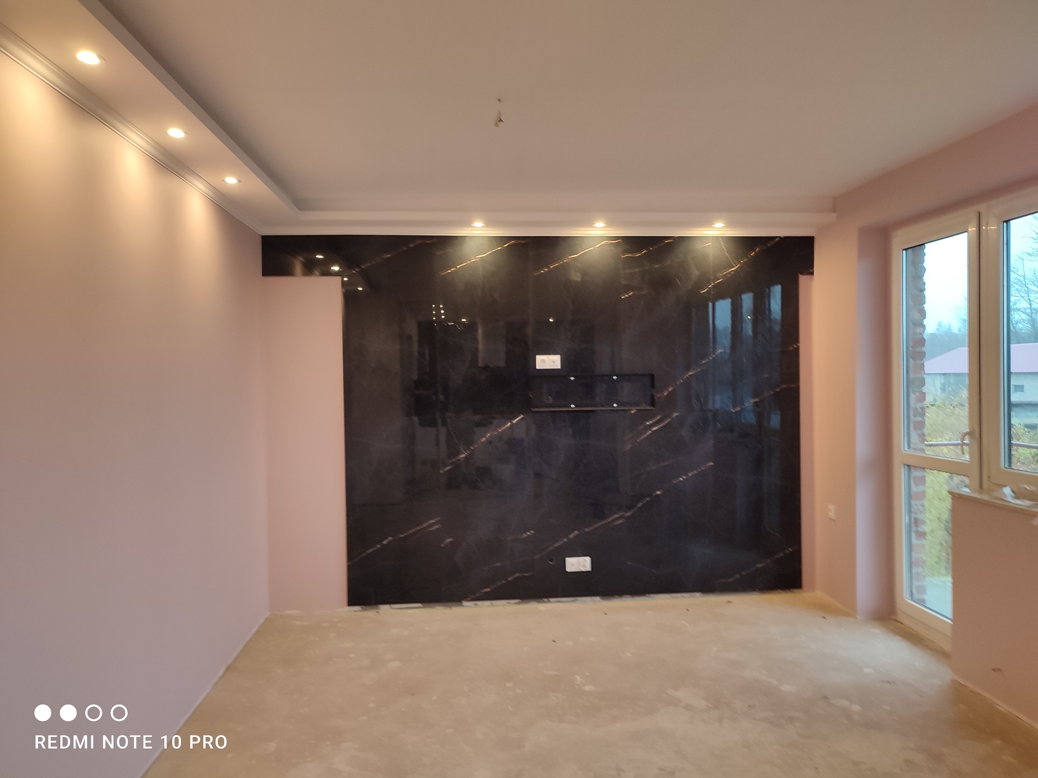 Wykończony pokój z czarną ścianą z efektem marmuru, oświetleniem punktowym na suficie i różowymi ścianami, podłoga przygotowana do dalszych prac wykończeniowych.