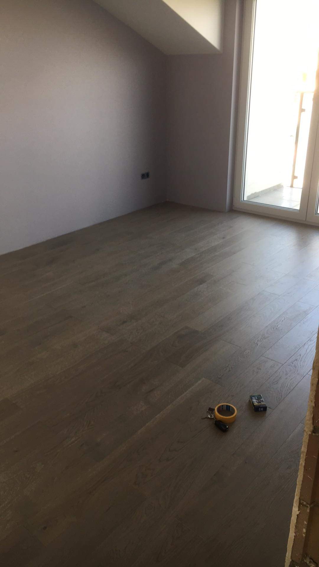 Jasne panele podłogowe ułożone w pokoju ze ścianami w odcieniach szarości, widoczne narzędzia i akcesoria do montażu podłogi, takie jak taśma klejąca i nóż.