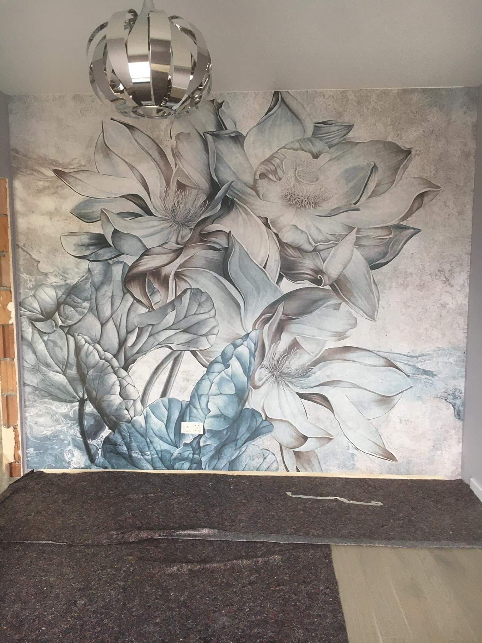 Ściana z fototapetą w kwiaty magnolii w odcieniach szarości i błękitu, zabezpieczona na podłodze grubą matą ochronną, z widocznym gniazdkiem elektrycznym i nowoczesną lampą sufitową.