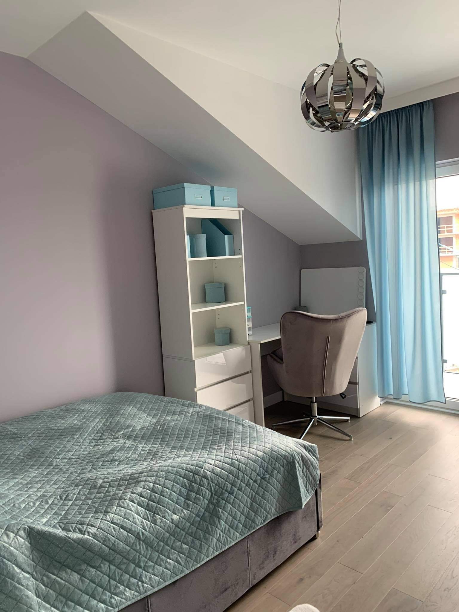 Pokój na poddaszu z łóżkiem, biurkiem i regałem w jasnych kolorach, oświetlony nowoczesną lampą wiszącą i światłem wpadającym przez błękitne zasłony.