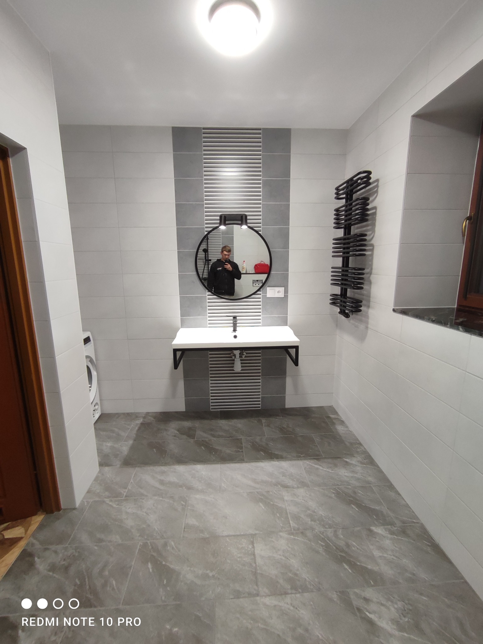 Wykończona łazienka z szarymi płytkami podłogowymi imitującymi marmur, biało-szare płytki ścienne, okrągłe lustro z podświetleniem i czarną ramą, biała umywalka na czarnych wspornikach oraz czarny...