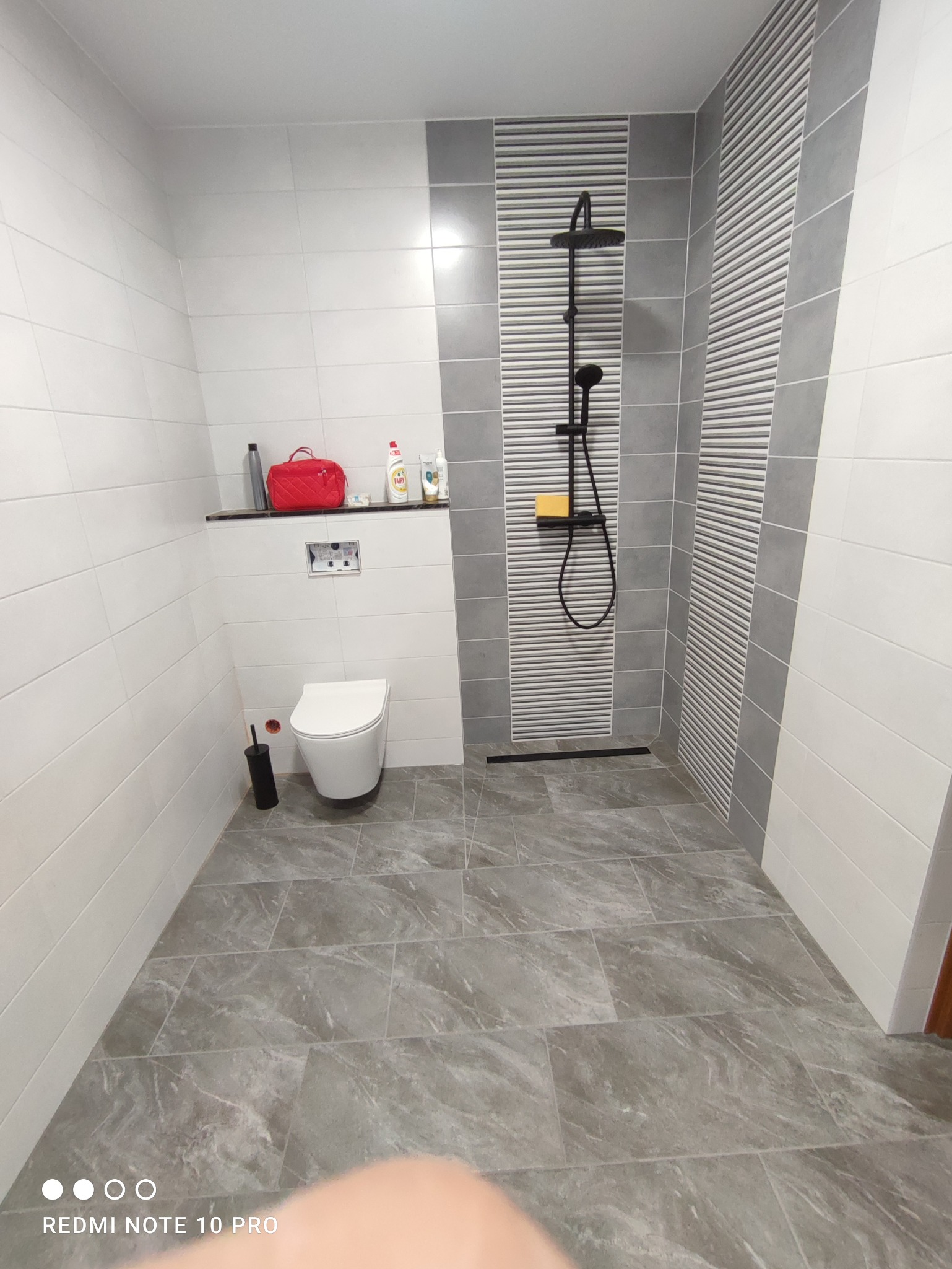 Wykończona łazienka z szarymi płytkami imitującymi marmur na podłodze, białe płytki na ścianach z szarymi pasami, czarna armatura prysznicowa i odpływ liniowy, toaleta podwieszana, półka...