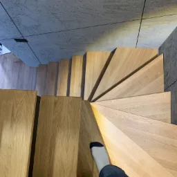 Widok z góry na spiralne schody z jasnego drewna, osoba w skarpetkach i jeansach schodzi w dół, szare płytki na ścianie.