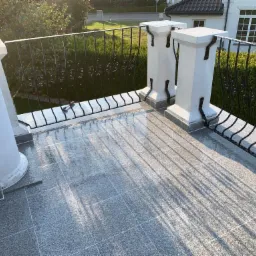 Wykończony balkon z szarą, kwadratową płytką podłogową, ozdobną, czarną metalową balustradą i białymi kolumnami w słoneczny dzień.