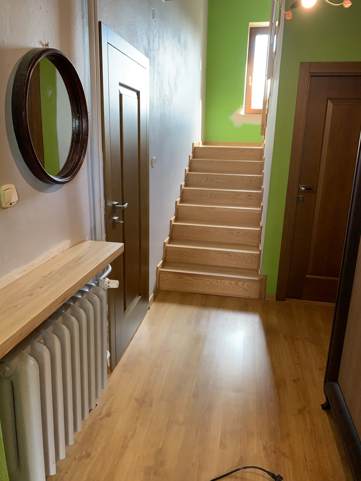 Jasne drewniane schody w korytarzu z zieloną ścianą, widok z dołu, obok drzwi i grzejnik z drewnianym blatem, na ścianie okrągłe lustro.