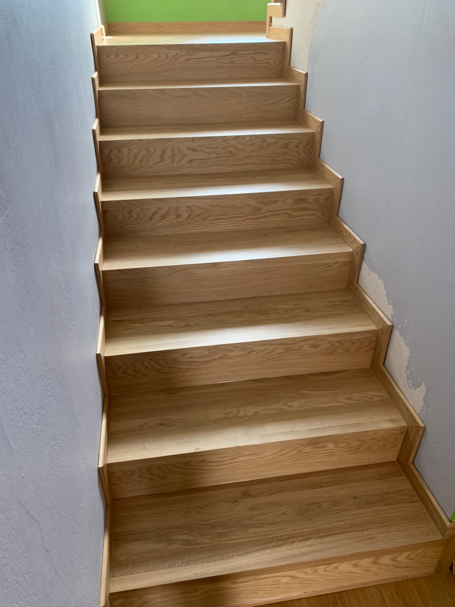 Nowe drewniane schody dębowe w trakcie montażu, widoczne surowe ściany w szarym kolorze i zielona ściana na górze, brak balustrady.