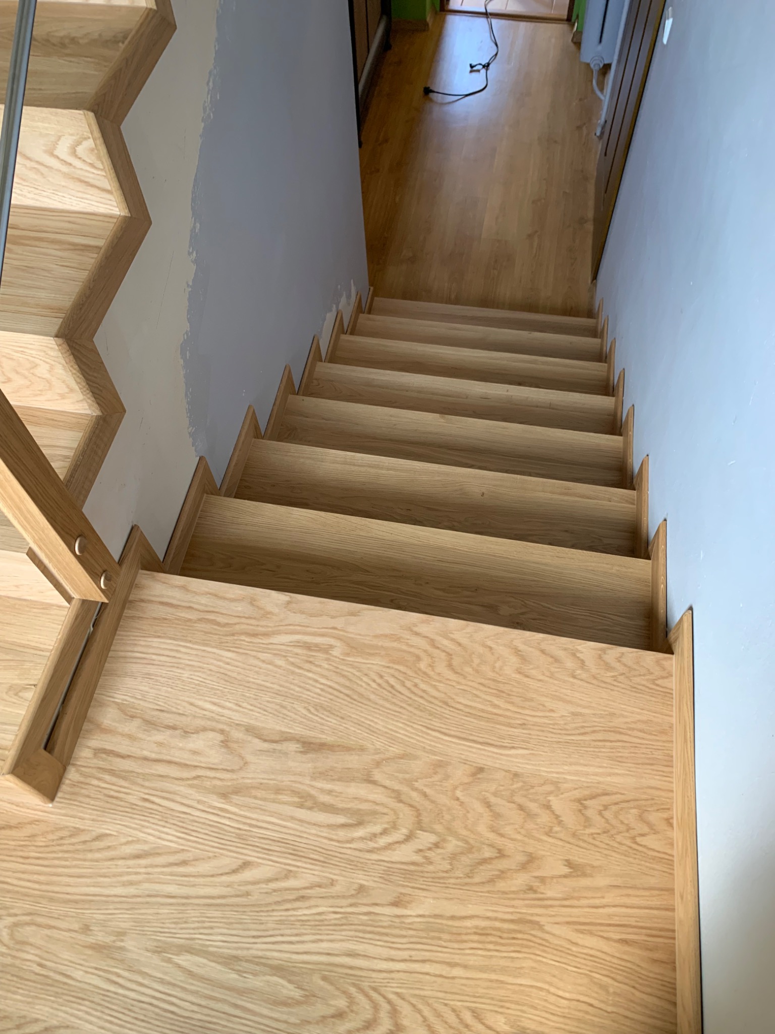 Widok z góry na jasne, dębowe schody z minimalistyczną balustradą, prowadzące w dół; ściana pomalowana na jasny szaro-niebieski kolor, widoczne niedoskonałości malowania przy łączeniu ze schodami.