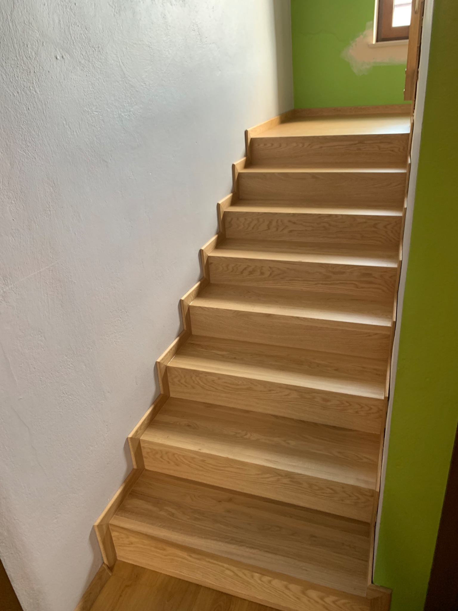 Nowe, jasne schody drewniane z litego drewna dębowego, widoczne z dołu, z zieloną ścianą po prawej i białą po lewej, prowadzące do góry.