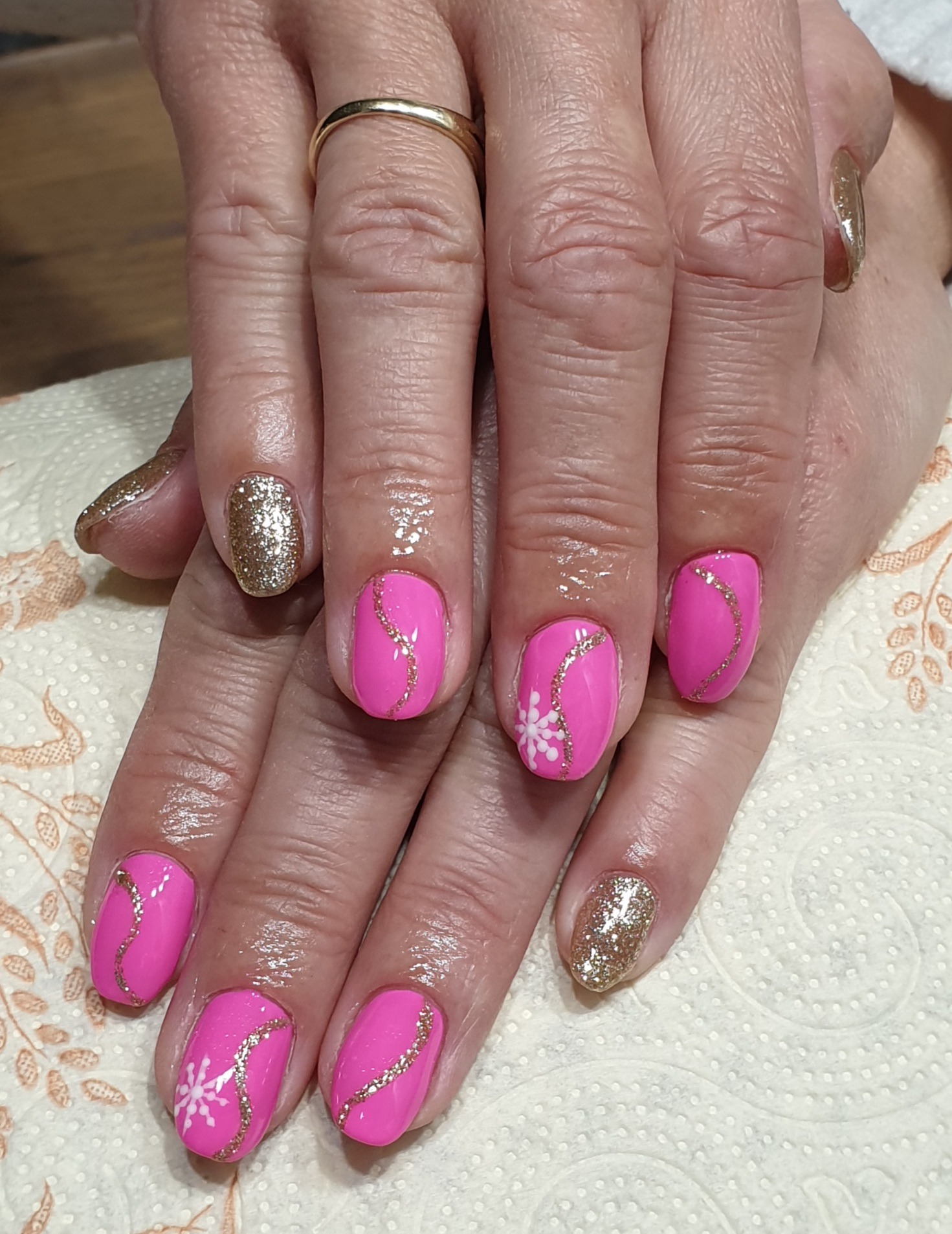 Ręce z manicure: różowe paznokcie z brokatową linią i motywem śnieżynki, dwa paznokcie w całości pokryte złotym brokatem, widoczny złoty pierścionek na palcu.