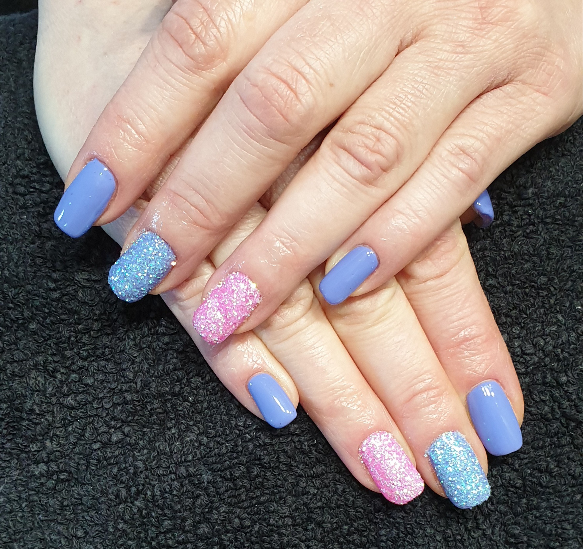 Dłonie z manicure: paznokcie pomalowane na fioletowo-niebieski kolor oraz dwa paznokcie pokryte brokatem w kolorach różowym i niebieskim na czarnym tle.