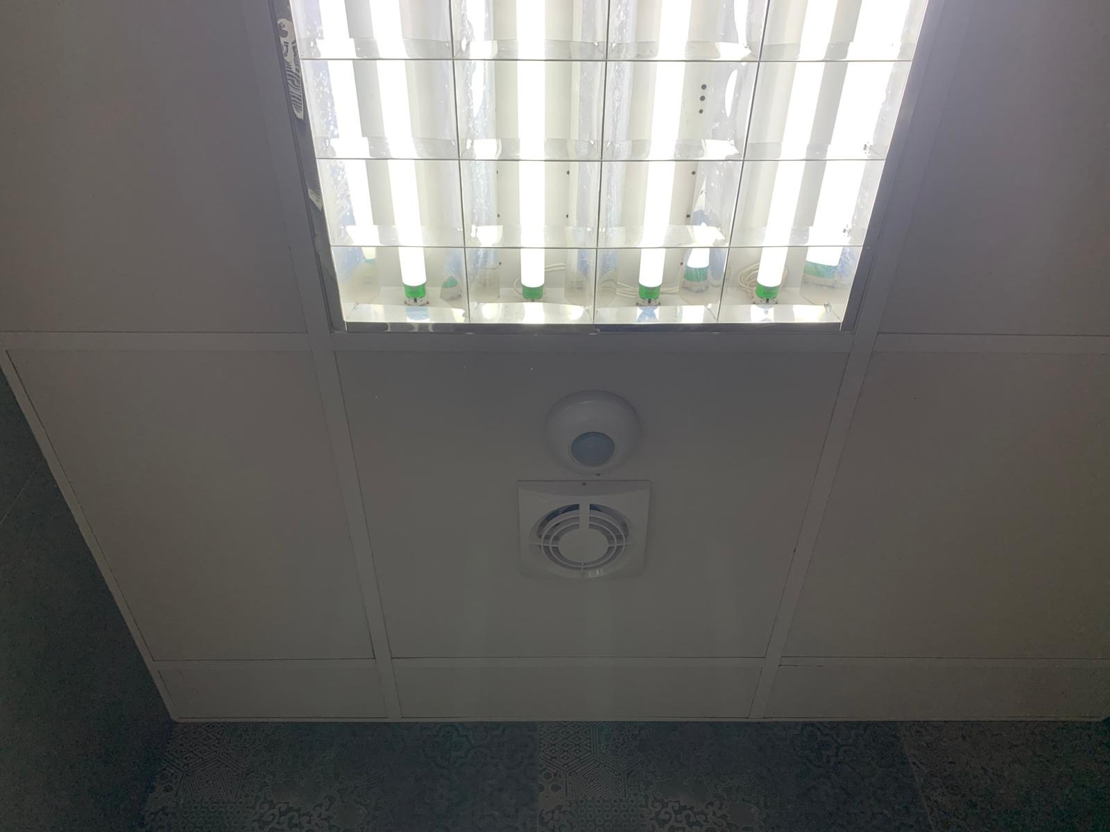 Sufit podwieszany z rastrową oprawą oświetleniową z lampami jarzeniowymi, czujnikiem ruchu i kratką wentylacyjną, widoczne płytki podłogowe w tle.