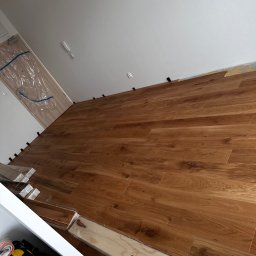Jakub Schlosberger Usługi Remontowe - Układanie paneli podłogowych w trakcie pracy, widoczne dystanse przy ścianie, paczki paneli w folii i skrzynka z narzędziami w rogu.