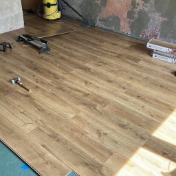 Jakub Schlosberger Usługi Remontowe - Układanie paneli podłogowych w pokoju, widoczne narzędzia: młotek gumowy, kątownik, piła do paneli, wkrętarka akumulatorowa oraz paczki paneli. Pod spodem zielony podkład i niebieska taśma. Ściany...