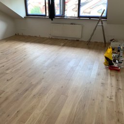 Jakub Schlosberger Usługi Remontowe - Jasne panele podłogowe w pokoju na poddaszu, widoczna drabina i narzędzia do wykończenia, listwy przypodłogowe jeszcze nie zamontowane.