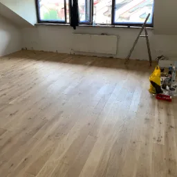 Jasne panele podłogowe w pokoju na poddaszu, widoczna drabina i narzędzia do wykończenia, listwy przypodłogowe jeszcze nie zamontowane.