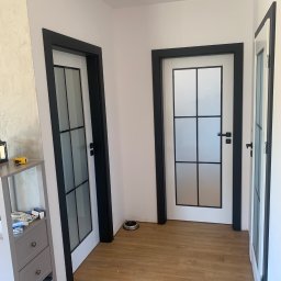 Jakub Schlosberger Usługi Remontowe - Trzy białe drzwi wewnętrzne z czarnymi ramami i szybami w stylu loft, zamontowane w korytarzu z panelami podłogowymi.