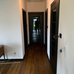 Jakub Schlosberger Usługi Remontowe - Korytarz w trakcie remontu z drewnianą podłogą, białymi ścianami z czarnymi listwami przypodłogowymi i czarnymi drzwiami. Widoczne niedokończone prace elektryczne przy jednym z otworów drzwiowych.