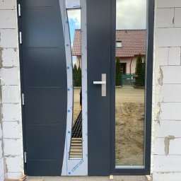 Jakub Schlosberger Usługi Remontowe - Nowe, grafitowe drzwi wejściowe z bocznym przeszkleniem, jeszcze z folią ochronną, zamontowane w surowym murze z białych bloczków; widoczne drewniane podkładki pod progiem.