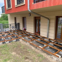 Jakub Schlosberger Usługi Remontowe - Konstrukcja drewnianego tarasu na betonowych bloczkach, czarna folia pod spodem, widok na budynek mieszkalny z oknami i balkonem.