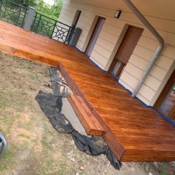 Jakub Schlosberger Usługi Remontowe - Nowo wybudowany, lakierowany drewniany taras przylegający do budynku z beżową elewacją, widoczne okna z brązowymi ramami, na ziemi leży czarna folia i stoi szara konewka.