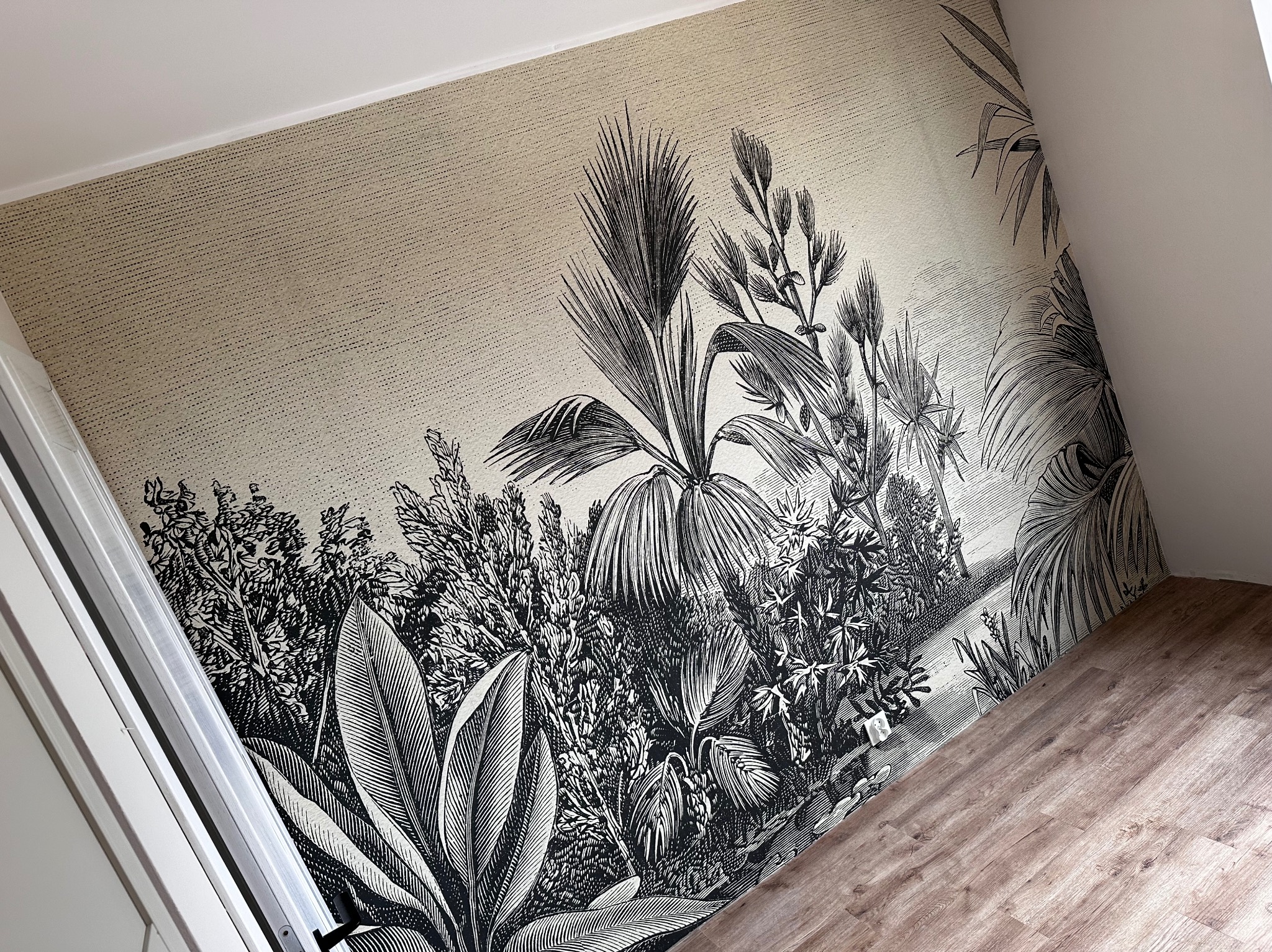 Ściana w pokoju na poddaszu pokryta beżową tapetą z czarnym, szczegółowym motywem roślin tropikalnych, widoczny fragment podłogi z paneli w odcieniach brązu.