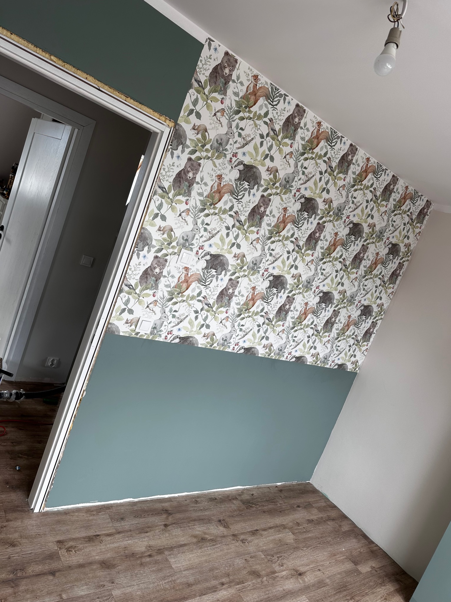 Ściana w pokoju, dolna część pomalowana na zielono, górna pokryta tapetą w leśne zwierzęta, widoczny fragment ościeżnicy drzwiowej i podłoga z paneli.