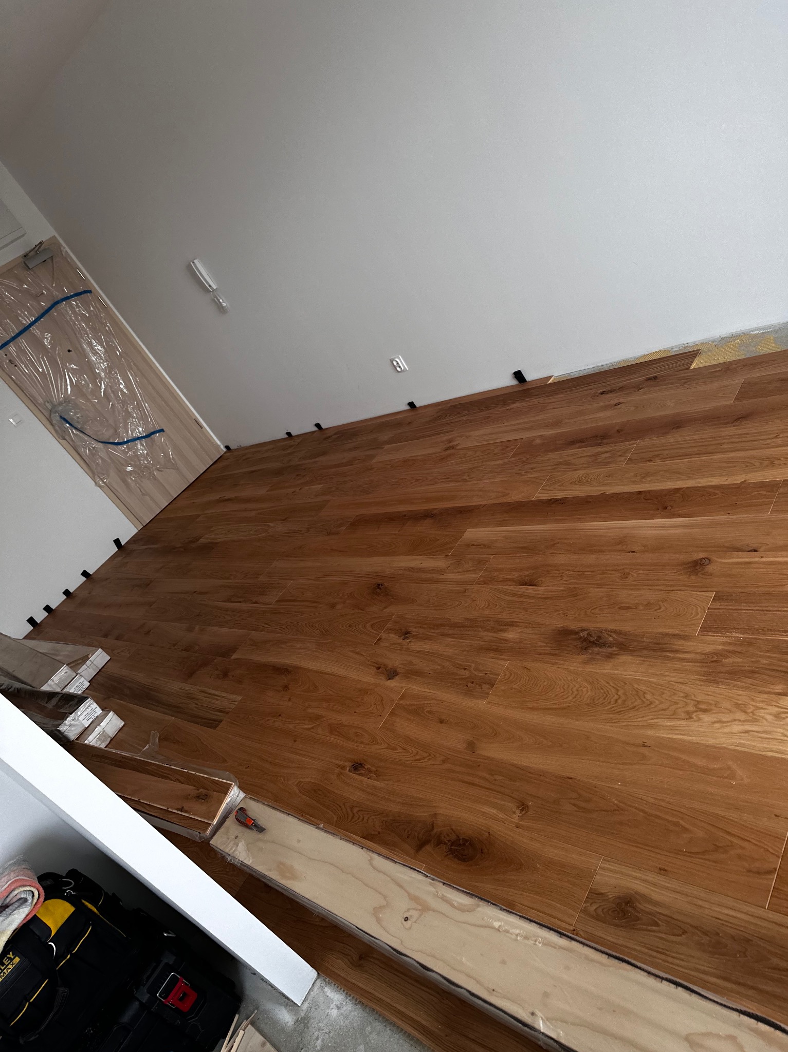 Układanie paneli podłogowych w trakcie pracy, widoczne dystanse przy ścianie, paczki paneli w folii i skrzynka z narzędziami w rogu.