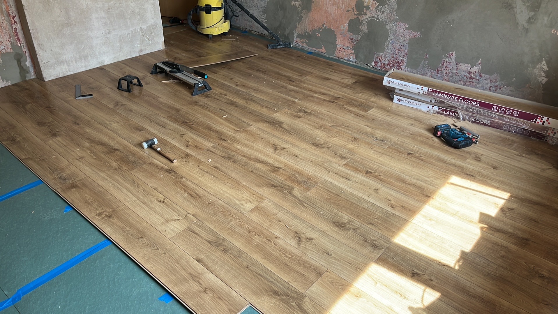 Układanie paneli podłogowych w pokoju, widoczne narzędzia: młotek gumowy, kątownik, piła do paneli, wkrętarka akumulatorowa oraz paczki paneli. Pod spodem zielony podkład i niebieska taśma. Ściany...