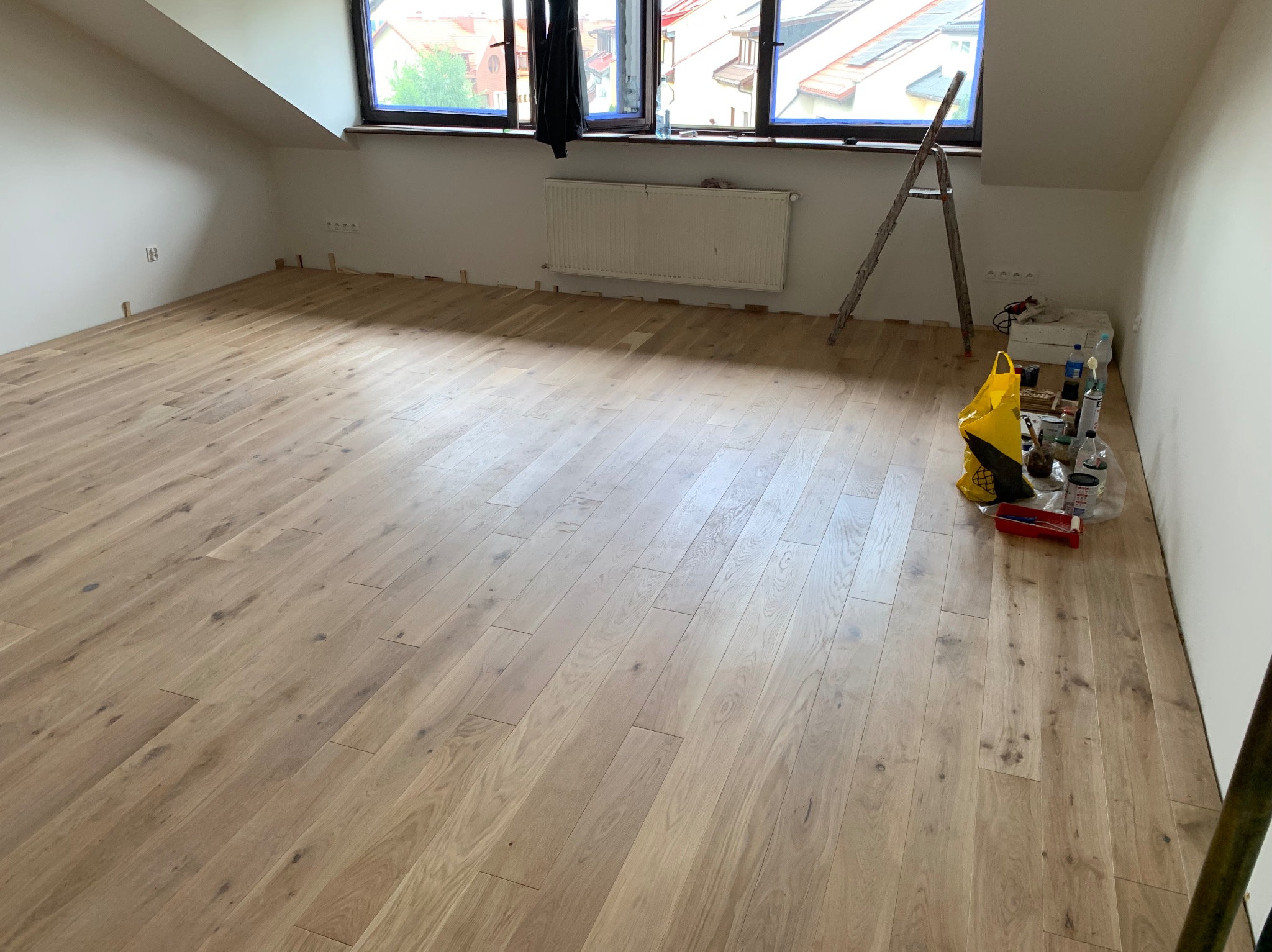 Jasne panele podłogowe w pokoju na poddaszu, widoczna drabina i narzędzia do wykończenia, listwy przypodłogowe jeszcze nie zamontowane.