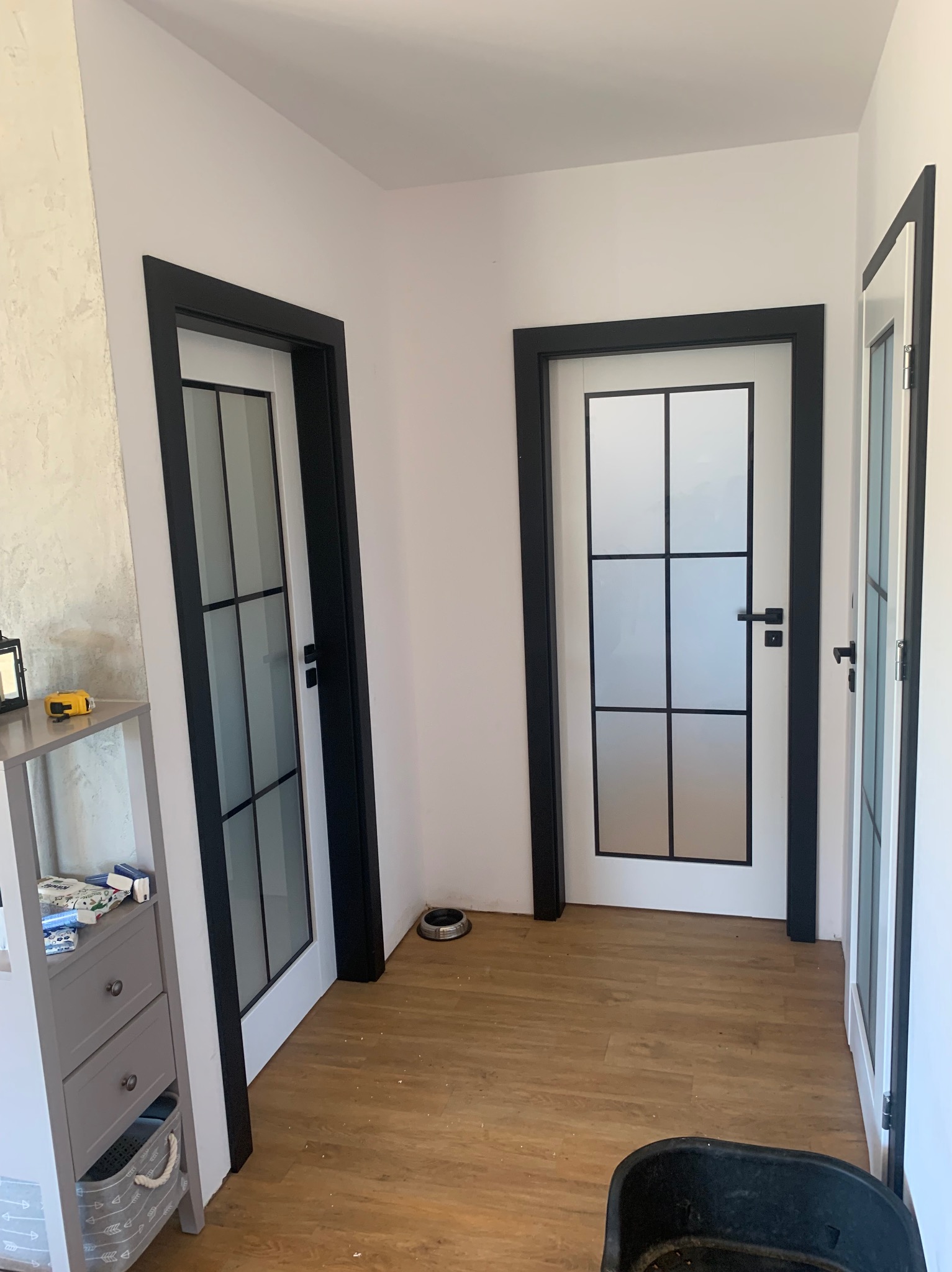 Trzy białe drzwi wewnętrzne z czarnymi ramami i szybami w stylu loft, zamontowane w korytarzu z panelami podłogowymi.