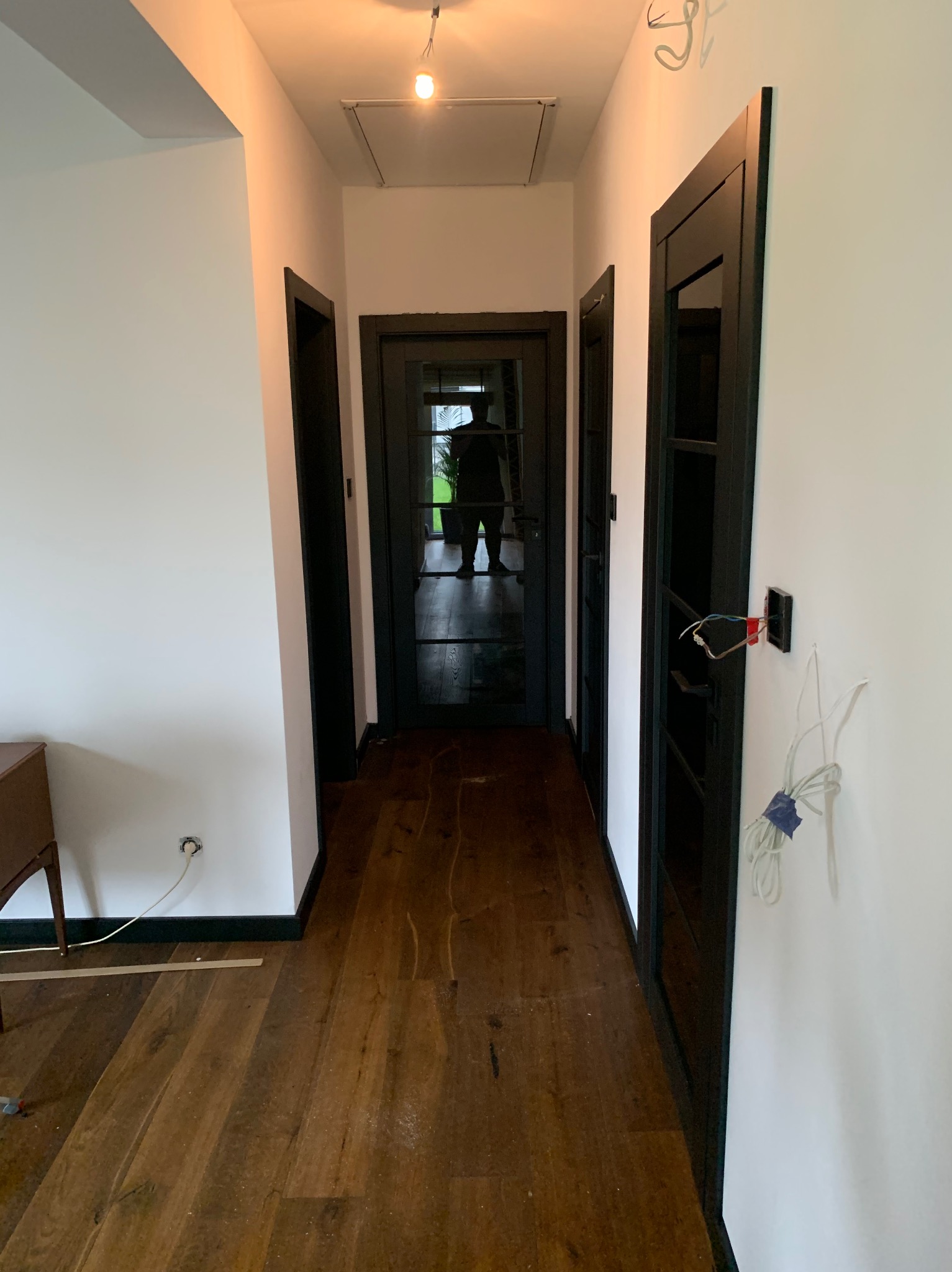 Korytarz w trakcie remontu z drewnianą podłogą, białymi ścianami z czarnymi listwami przypodłogowymi i czarnymi drzwiami. Widoczne niedokończone prace elektryczne przy jednym z otworów drzwiowych.