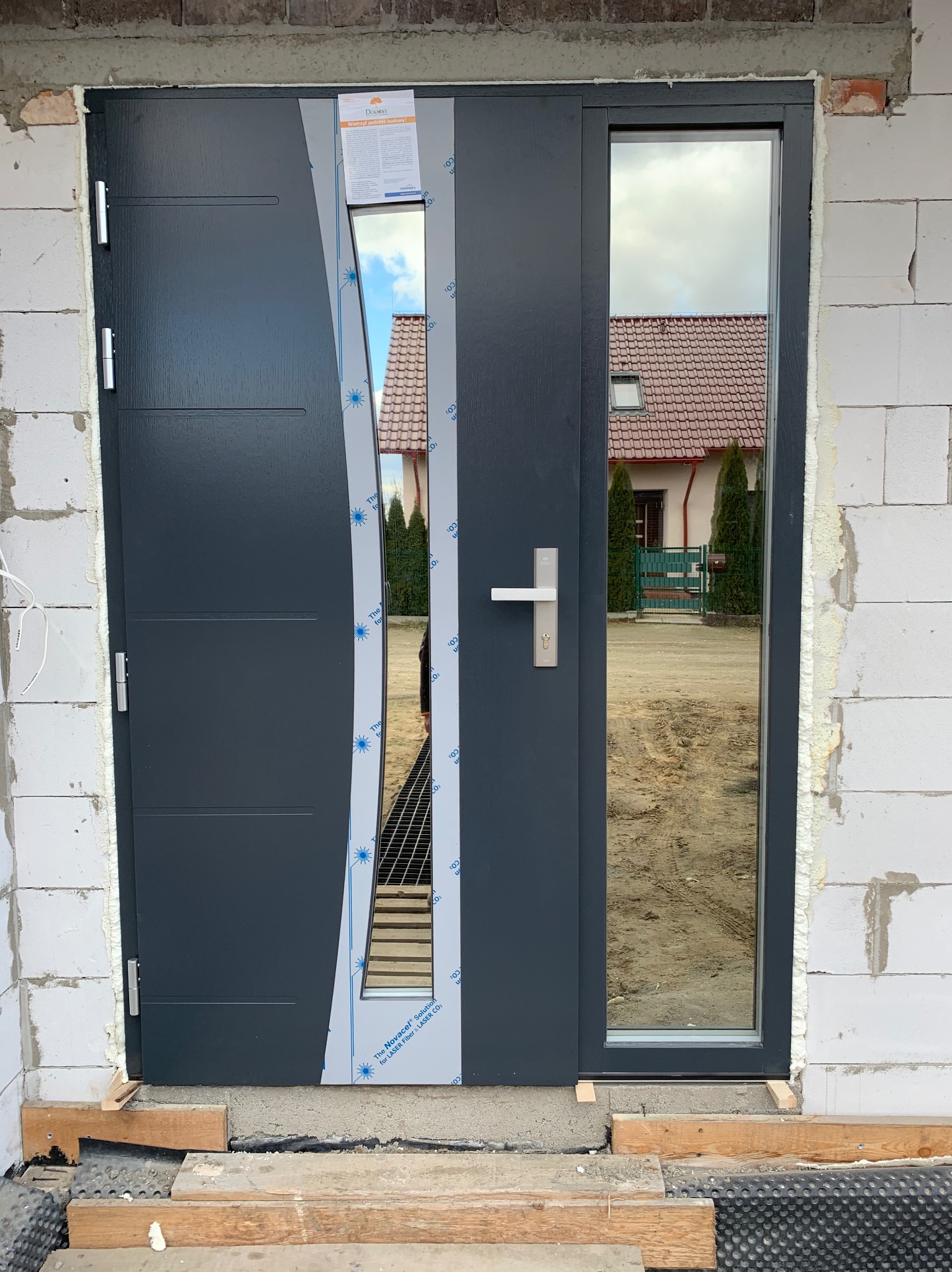 Nowe, grafitowe drzwi wejściowe z bocznym przeszkleniem, jeszcze z folią ochronną, zamontowane w surowym murze z białych bloczków; widoczne drewniane podkładki pod progiem.