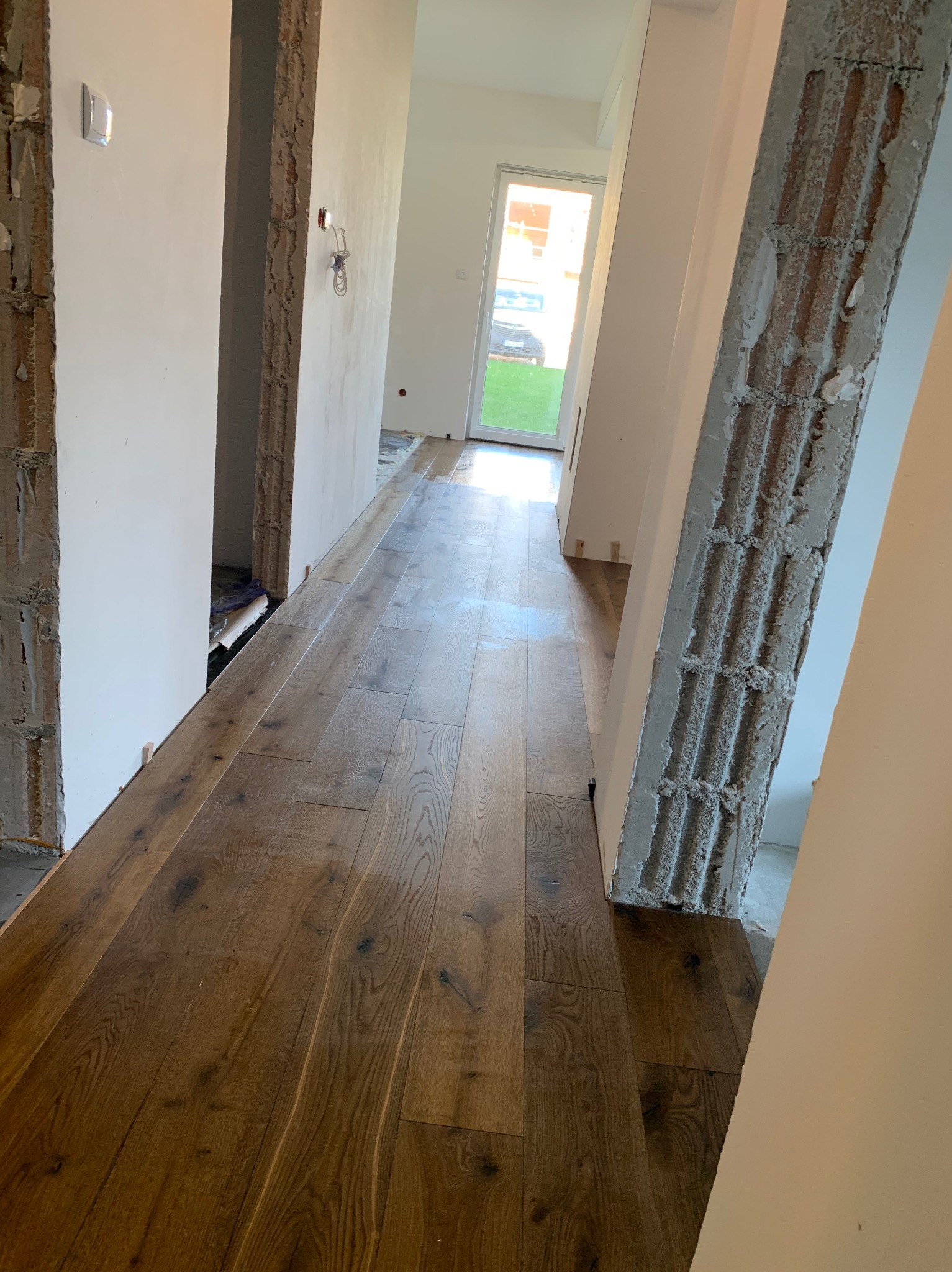 Korytarz w trakcie remontu z nowo ułożonymi panelami podłogowymi o ciepłym, brązowym odcieniu; widoczne surowe ściany z odsłoniętym betonem i instalacjami, oraz białe, pomalowane ściany. Listwy...