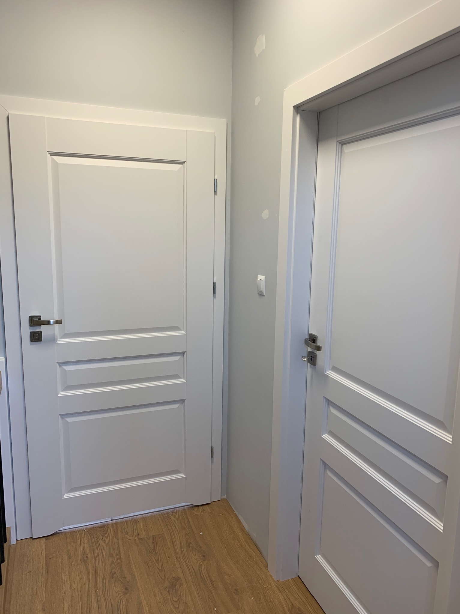 Dwa białe, klasyczne drzwi wewnętrzne z srebrnymi klamkami zamontowane w mieszkaniu w Trzebnicy, widoczne nierówności na ścianie wymagające poprawek.