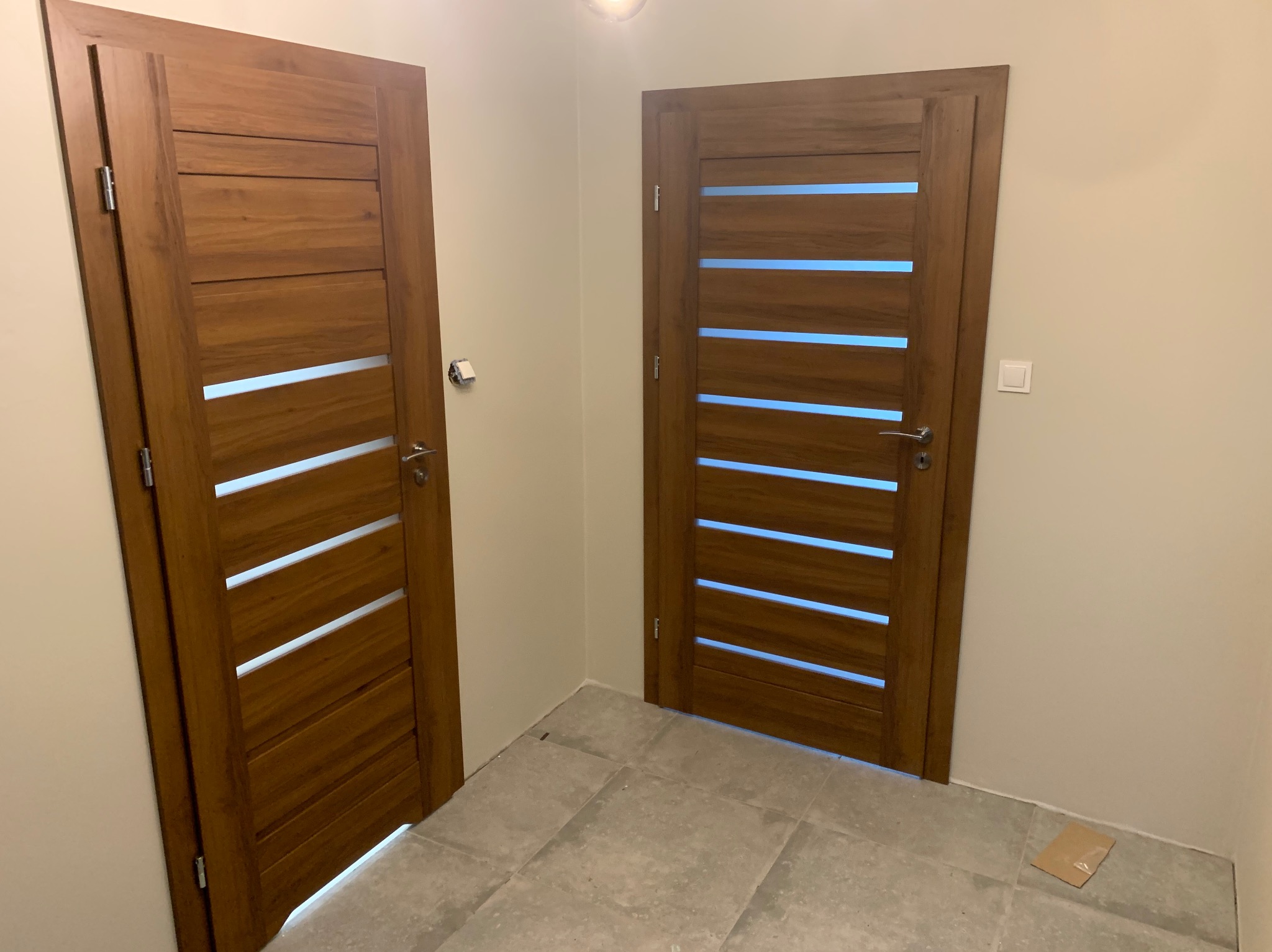 Dwie drewniane drzwi wewnętrzne z poziomymi przeszkleniami, zamontowane w jasnym korytarzu, widoczne na tle szarej podłogi z płytek.