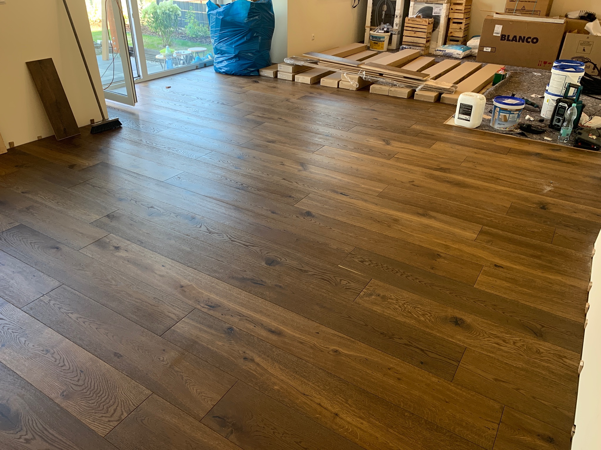 Ułożone panele podłogowe z widocznymi narzędziami, opakowaniami i workiem budowlanym w tle, częściowo otwarte drzwi balkonowe.