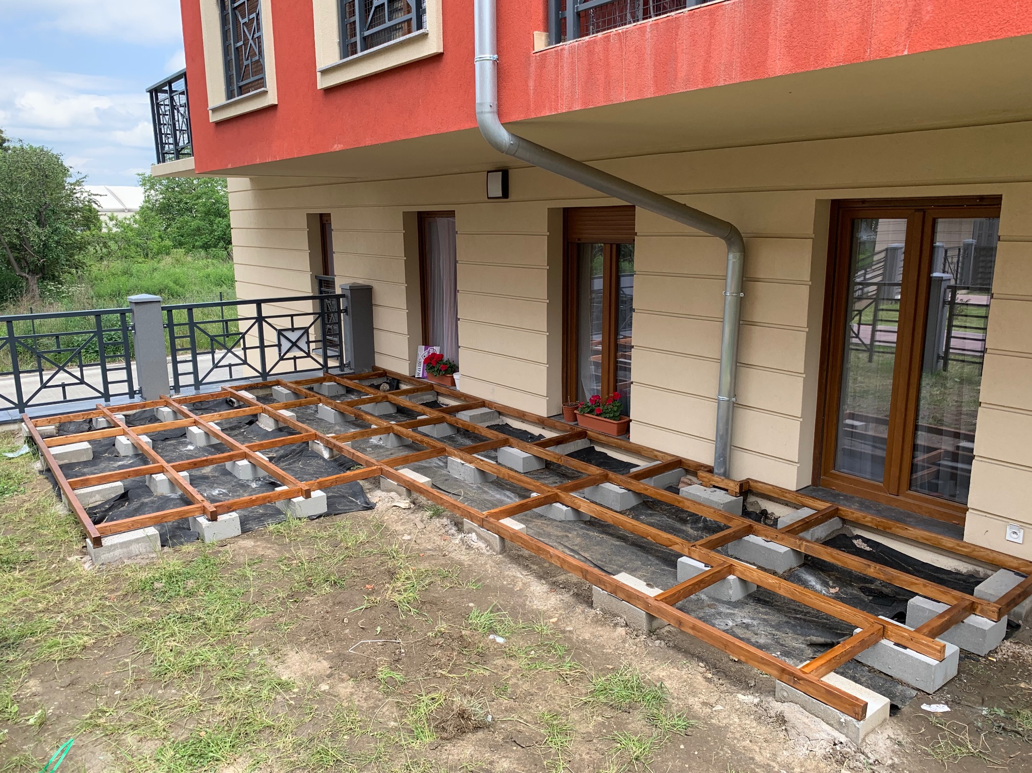 Konstrukcja drewnianego tarasu na betonowych bloczkach, czarna folia pod spodem, widok na budynek mieszkalny z oknami i balkonem.