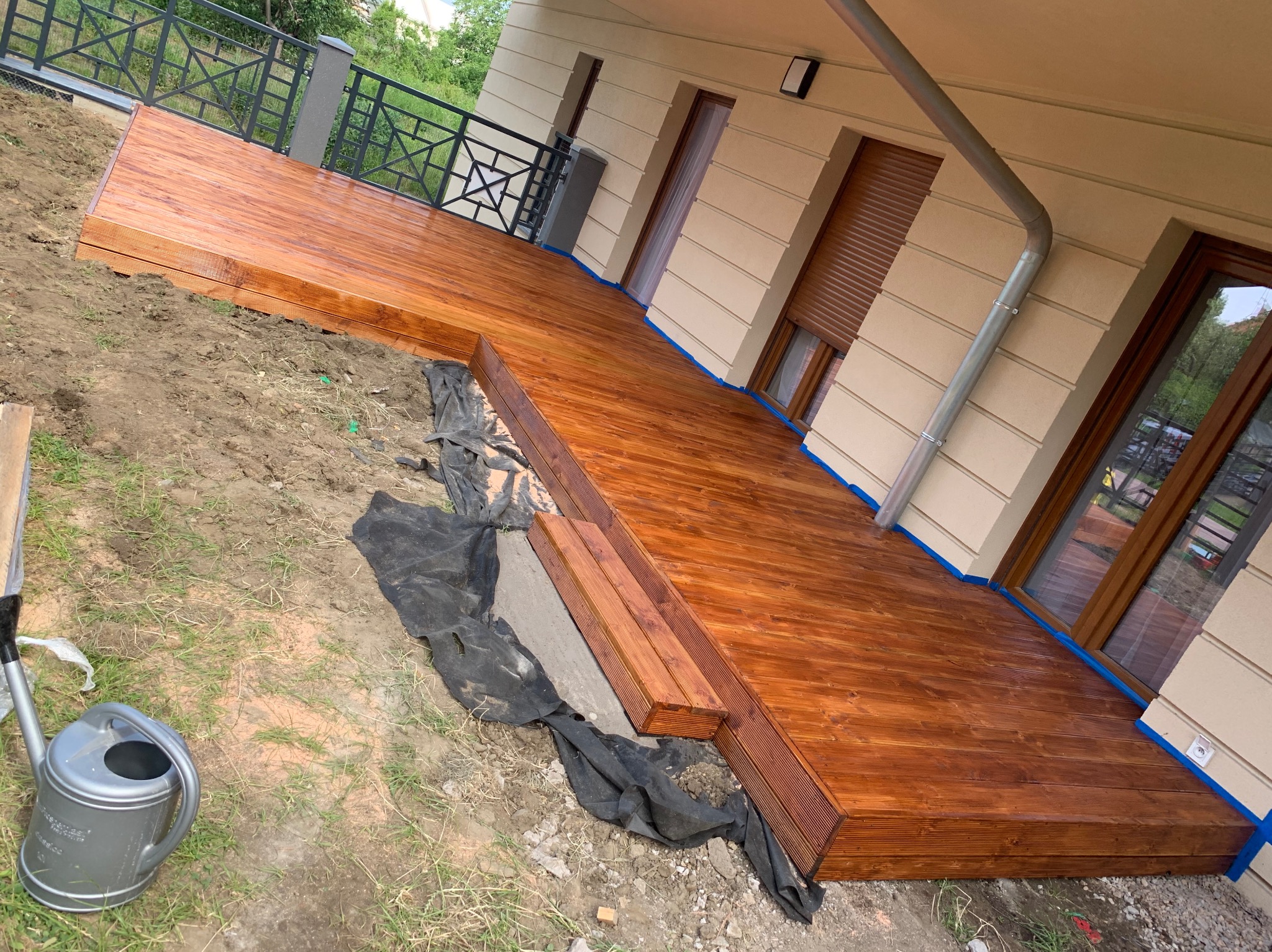 Nowo wybudowany, lakierowany drewniany taras przylegający do budynku z beżową elewacją, widoczne okna z brązowymi ramami, na ziemi leży czarna folia i stoi szara konewka.