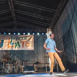 Uśmiechnięty mężczyzna w okularach i muszce, w jasnej koszuli i ż&oacute;łtych spodniach, tańczy na scenie podczas wydarzenia plenerowego 'Lubickie Lato', w tle widoczna perkusja i sprzęt nagłośnieniowy.