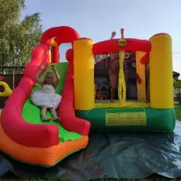 Dziewczynka w białej sukience z uśmiechem zjeżdża ze zjeżdżalni dmuchanego zamku Happy Hop, druga osoba w dmuchanym zamku, zielona trawa i niebieskie niebo w tle.