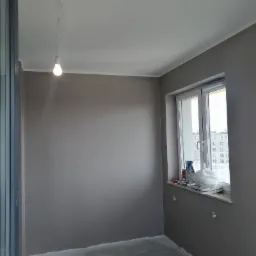 Pokój po szpachlowaniu i malowaniu na szaro, widoczne okno z białymi ramami, biały sufit z wiszącą żarówką, resztki materiałów budowlanych na parapecie okiennym.