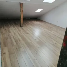 Poddasze po remoncie z drewnopodobną podłogą, drewnianą belką konstrukcyjną, białymi ścianami i oknami dachowymi.